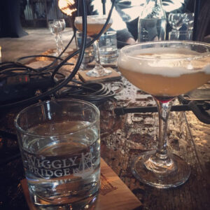 a table filled with bottles and glasses, a wiggly bridge glass on the coaster and an orange cocktail in a tall glass