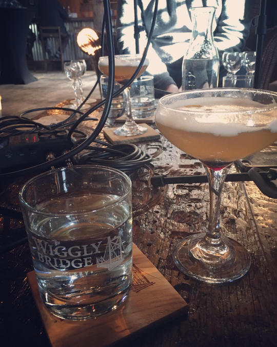 a table filled with bottles and glasses, a wiggly bridge glass on the coaster and an orange cocktail in a tall glass