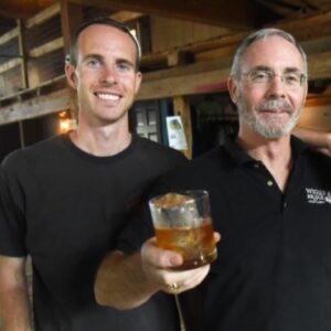 two men standing and posing, the man on the right is holding a glass out in a "cheers" manner